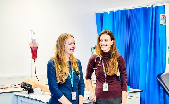 Two-people-standing-in-a-clinical-laboratory-wearing-stethoscopes-in-front-of-a-blue-curtain.